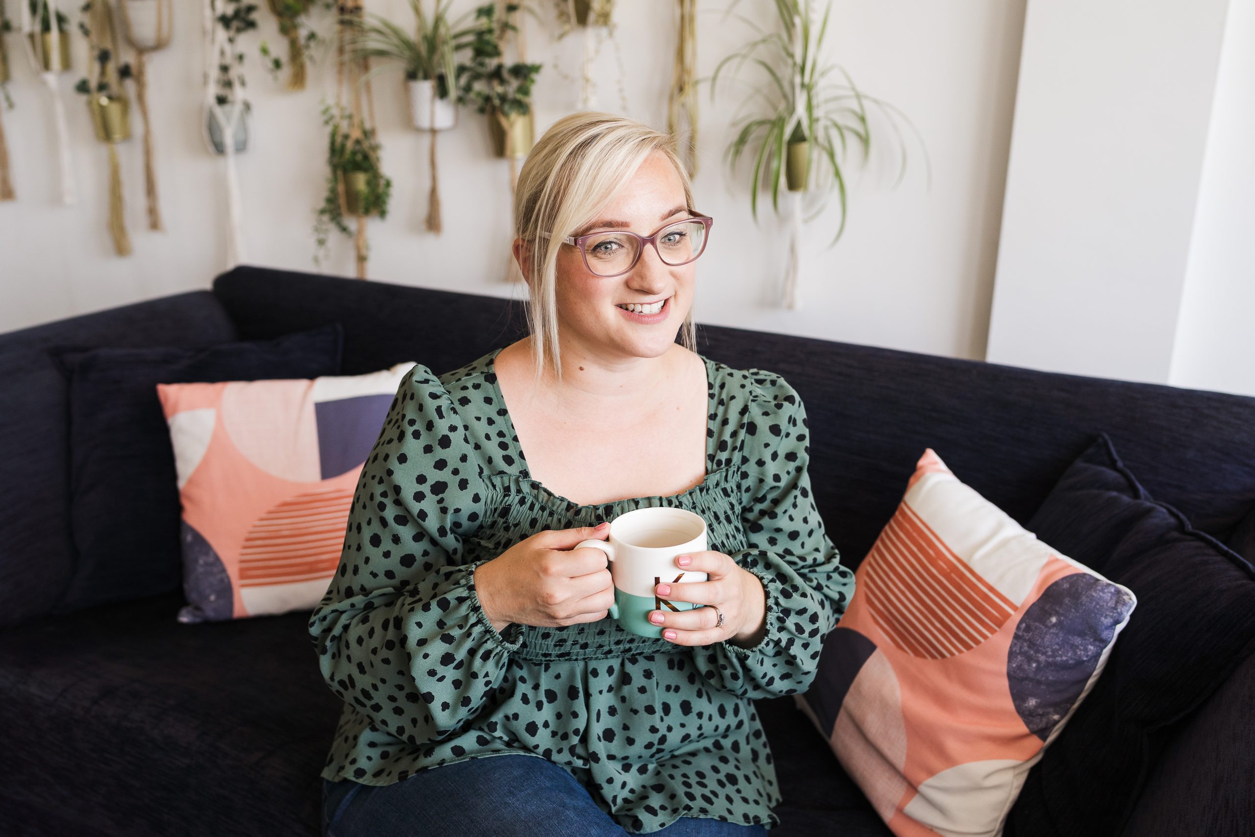 Katie, a white woman with blonde hair and glasses sat on a sofa with a cup of tea.