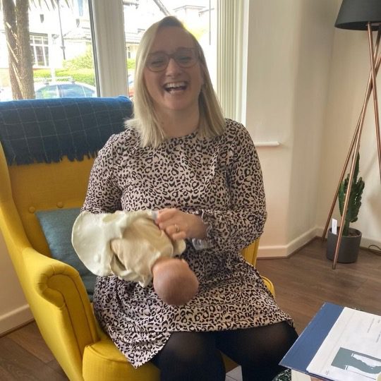 Katie, a white, blonde woman with pink glasses, holds a baby and pelvis and laughs with her clients.
