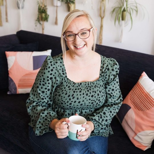 Katie, a white blonde woman with pink glasses and a green top with black spots, sits on a sofa with a cup of tea, smiling at the camera.